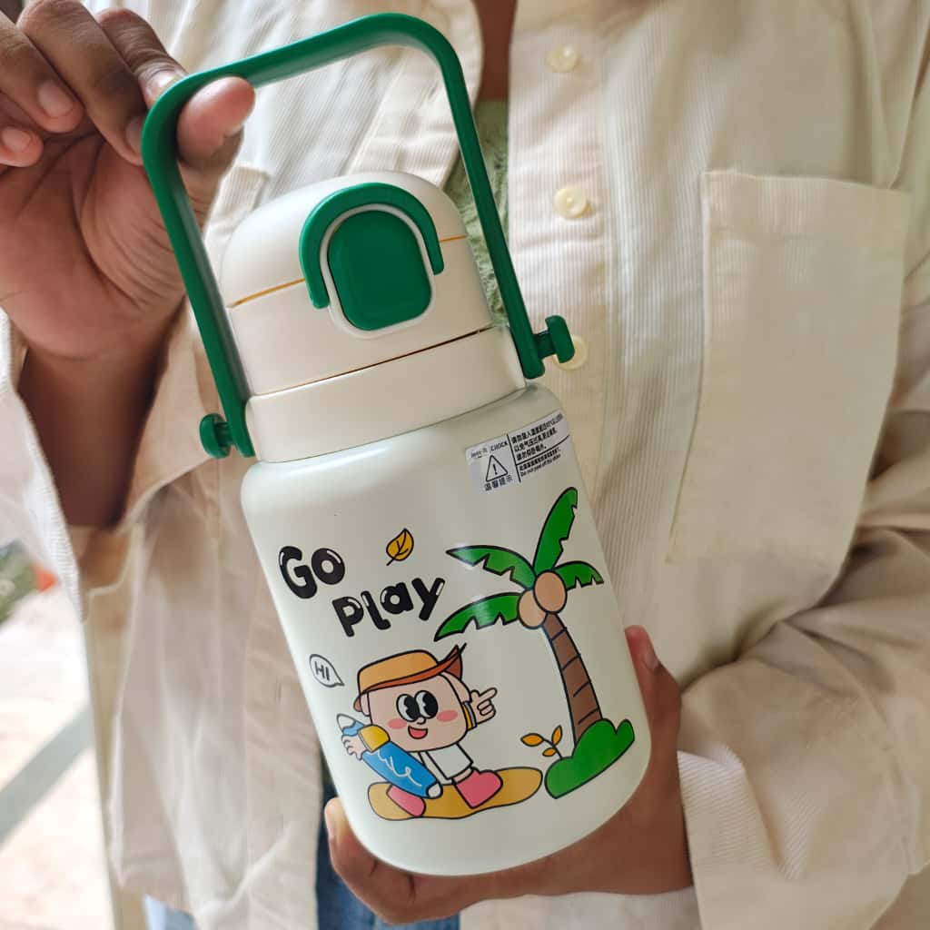 Person holding a white thermos with green handle and cartoon design, text 'Go Play' on a blurred background.