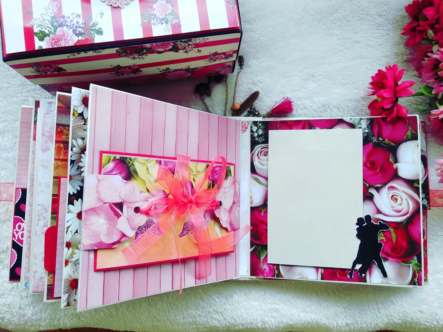 An open handmade paper album or scrapbook with pink floral covers, layered paper tabs, and a blank white photo slot on the right page, displayed on a white textured surface with decorative flowers.