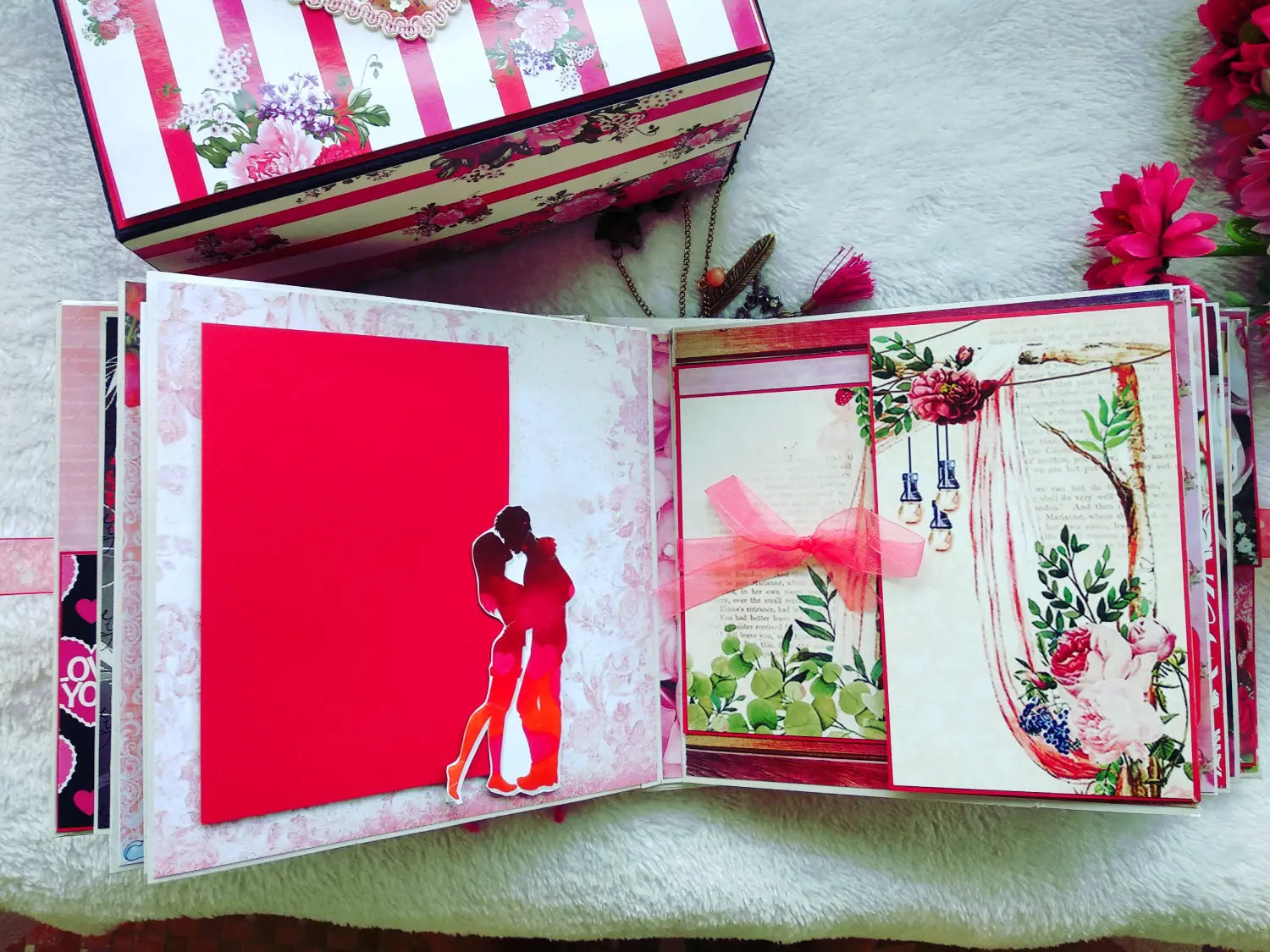 An open handmade paper album or scrapbook book displayed on a soft white surface, with a matching floral-patterned box partially visible above it. The pages feature decorative pink and red elements, including a large red panel with a couple silhouette and floral collage artwork.