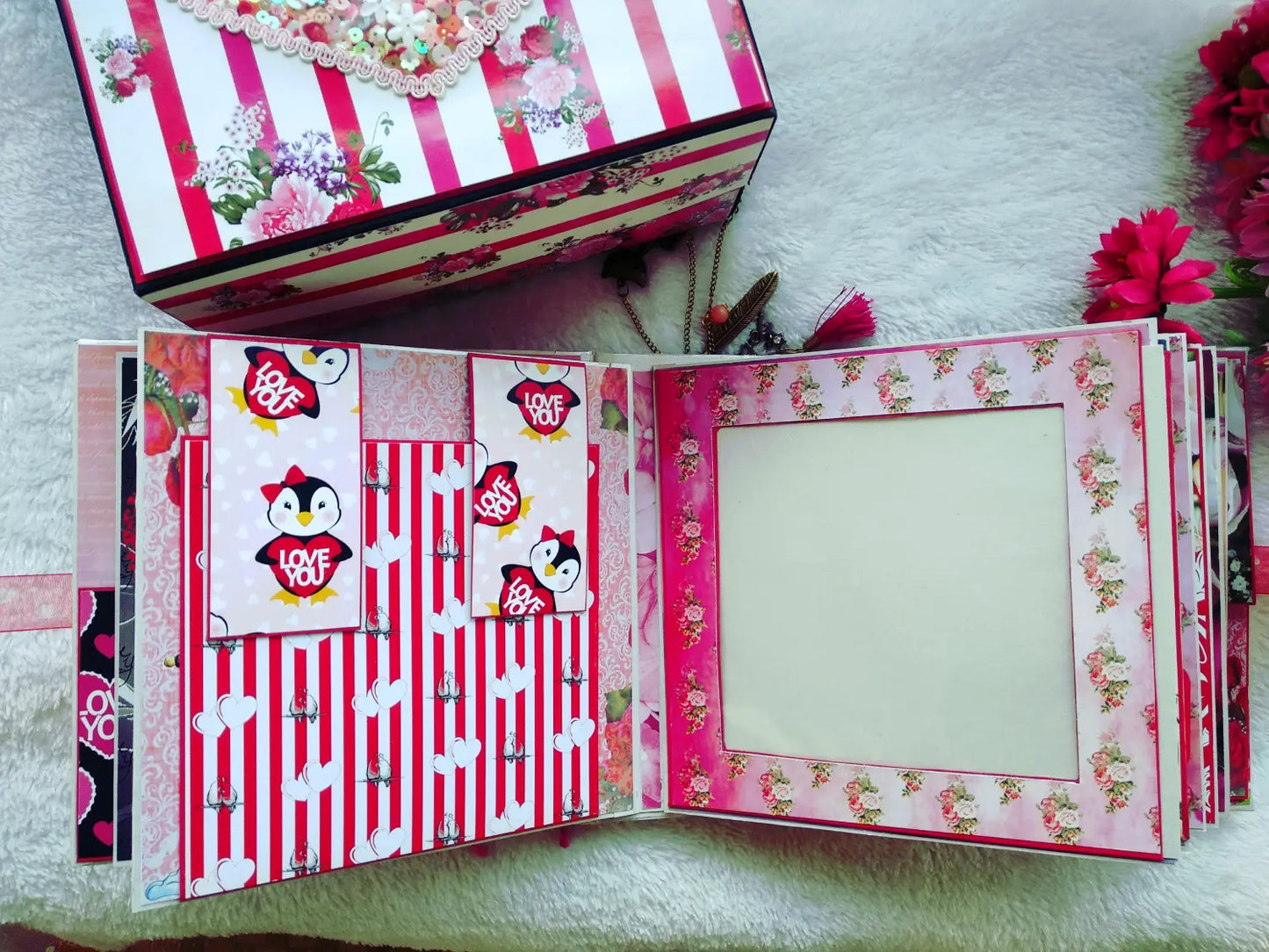 A handmade pink and red scrapbook album shown open, featuring penguin stickers, striped patterns, and floral borders, with a matching decorated box placed above it on a white textured surface.