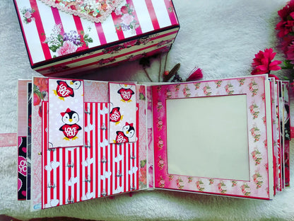 A handmade pink and red scrapbook album shown open, featuring penguin stickers, striped patterns, and floral borders, with a matching decorated box placed above it on a white textured surface.