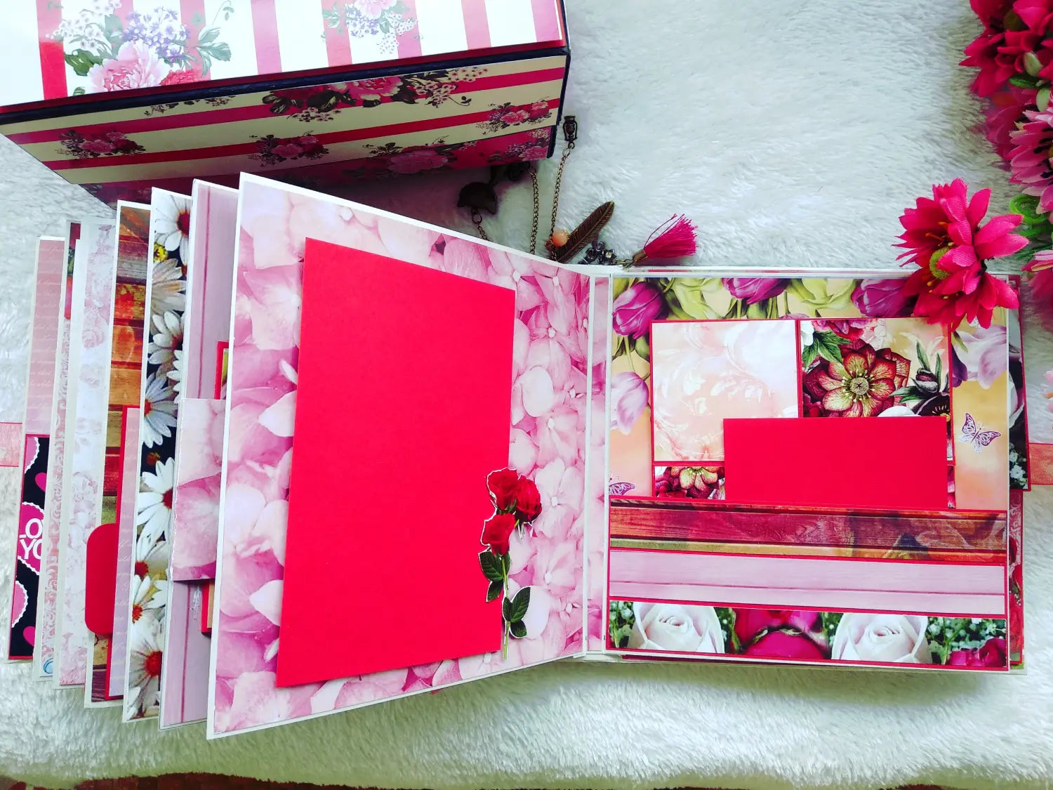 A handmade, multi-layered scrapbook album or memory book shown open, featuring pink floral patterns, red paper inserts, and decorative flowers on a white textured blanket.