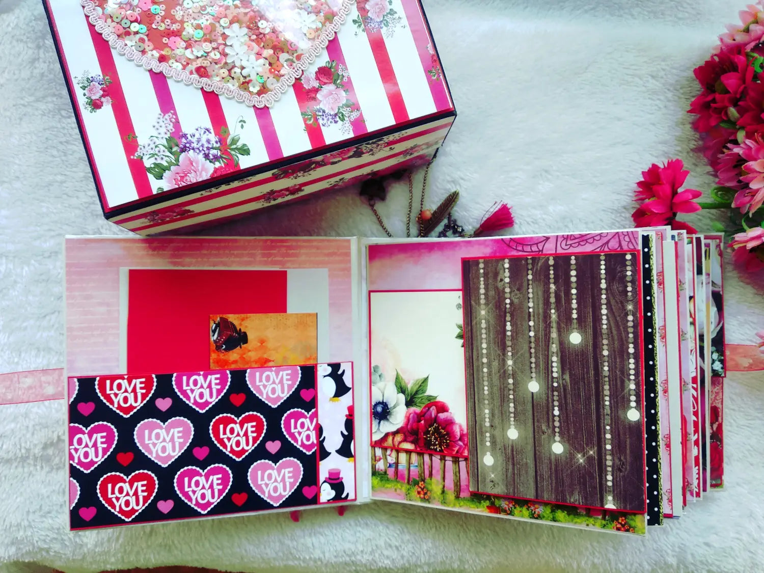 A handmade Valentine's Day scrapbook album and matching gift box displayed on a white textured blanket with decorative flowers. The album features multiple pages with pink and red patterns, including hearts, stripes, and floral designs, and is shown both closed on its box and open to display its interior pages.
