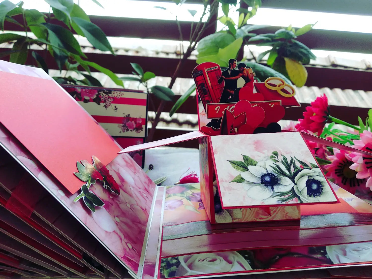 A close-up of a red and pink pop-up greeting card or gift box with floral artwork, shown with other matching card pieces and decorative flowers in a styled setting.