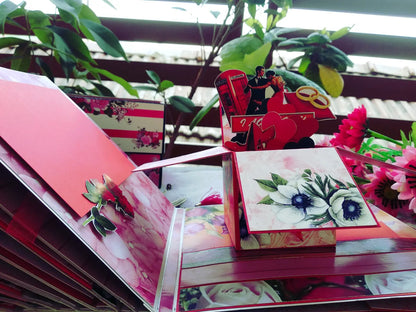 A close-up of a red and pink pop-up greeting card or gift box with floral artwork, shown with other matching card pieces and decorative flowers in a styled setting.