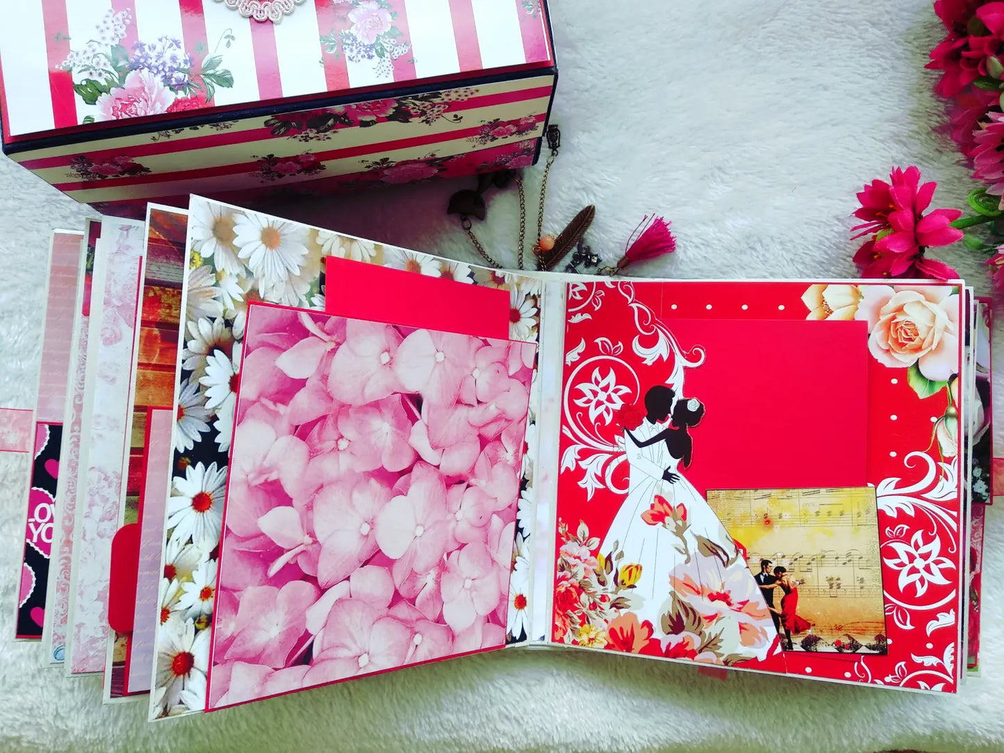 An open handmade paper album or scrapbook book displayed on a white textured surface, showing decorated inner pages with pink flowers, red patterns, floral borders, and a small photo insert. A matching floral box is partially visible at the top left.