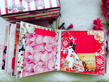 An open handmade paper album or scrapbook book displayed on a white textured surface, showing decorated inner pages with pink flowers, red patterns, floral borders, and a small photo insert. A matching floral box is partially visible at the top left.