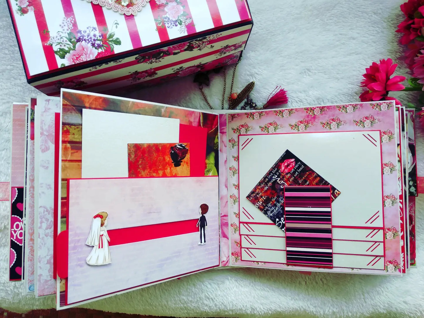 A handmade scrapbook album shown open, featuring pink and white striped pages, a house-shaped collage, and a bride-and-groom paper cutout. A matching floral and striped keepsake box is partially visible at the top, placed on a white textured surface with decorative flowers around it.
