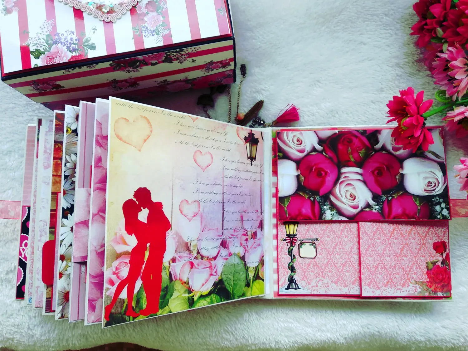 An open handmade scrapbook album or gift book displayed on a white textured surface, with a matching patterned gift box partially visible behind it. The pages feature floral designs, pink roses, heart motifs, a red couple silhouette, and decorative paper panels.