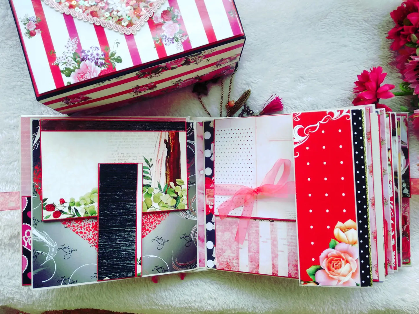 A handmade paper scrapbook album shown open, featuring multiple decorated pages in red, white, black, and floral colors, with a matching patterned storage box and artificial flowers on a white textured background.