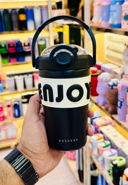 Black and white tumbler with 'ENJOY' text held in a hand against a colorful store background