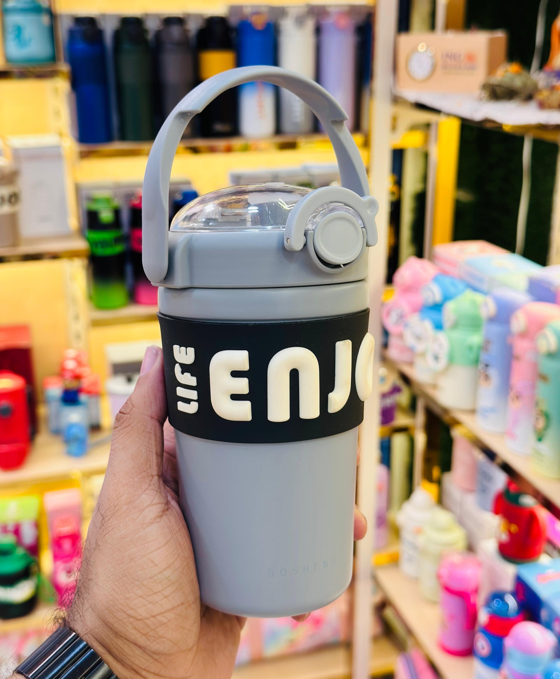 Gray water bottle with 'LIFE ENJOY' band held in a hand against a store background