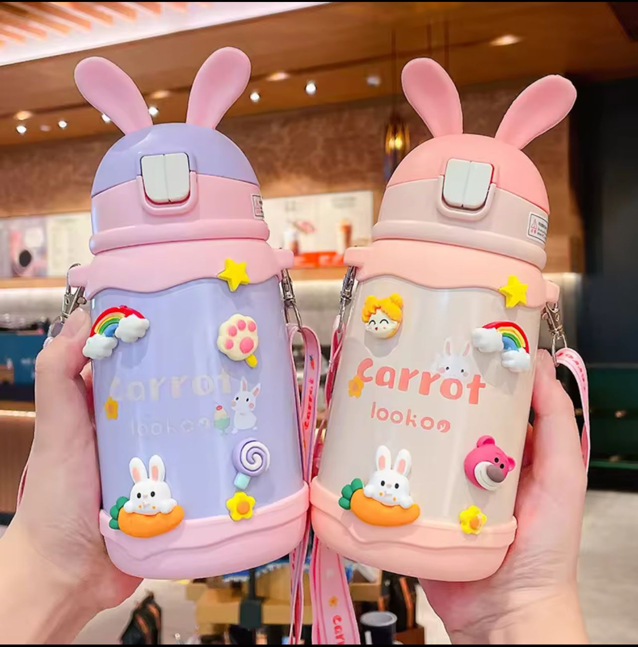 Two cute water bottles with bunny designs held in hands, one purple and one pink, against a blurred indoor background.