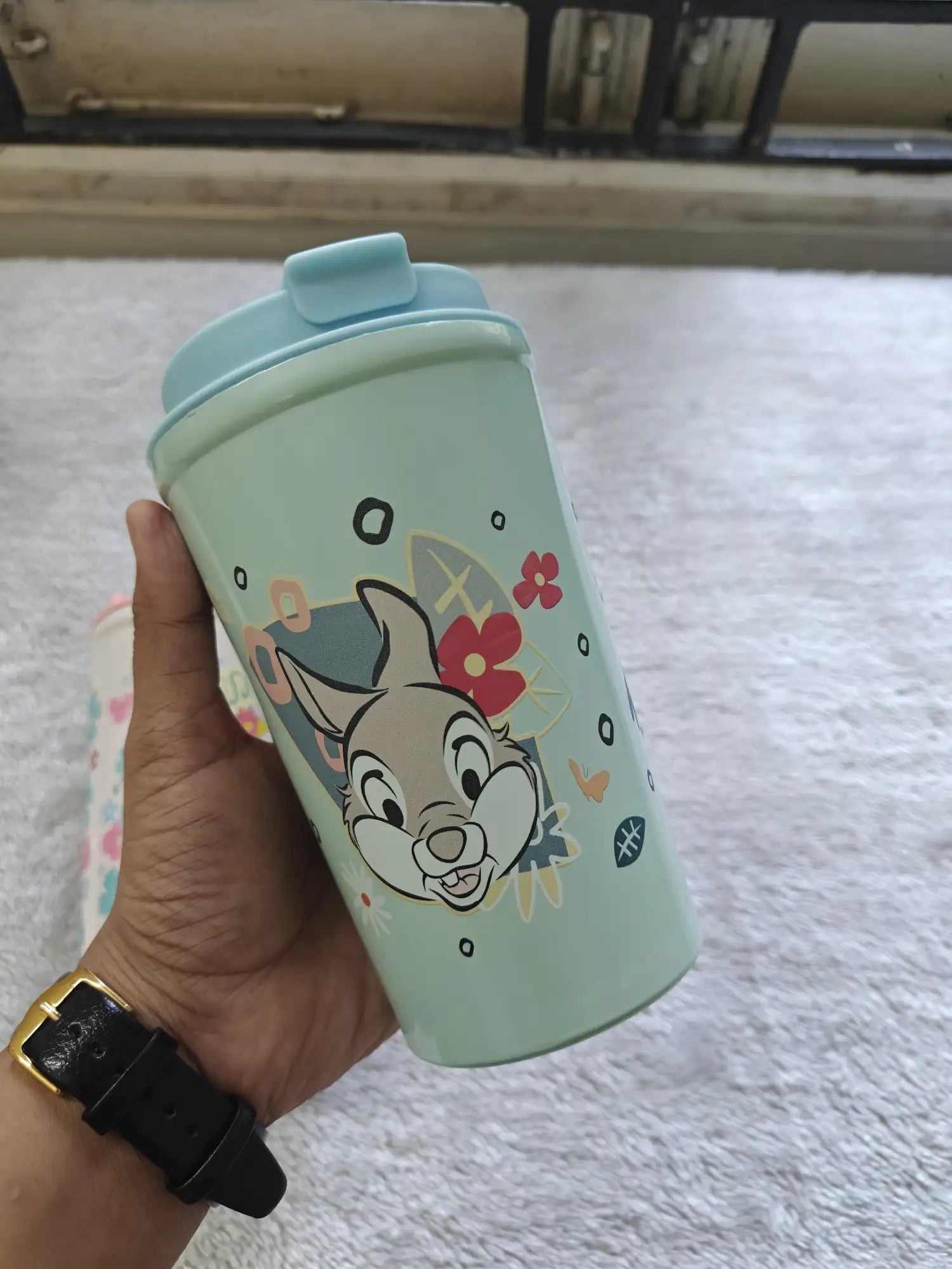 Hand holding a green tumbler with a cartoon character design on a light gray surface.