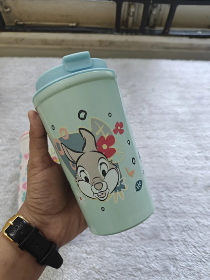 Hand holding a green tumbler with a cartoon character design on a light gray surface.