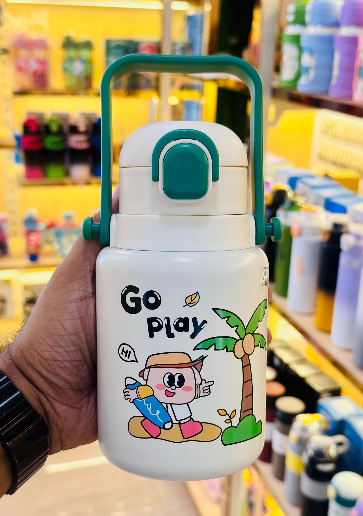 White thermos with green handle and 'Go Play' design held in a hand against a blurred store background.