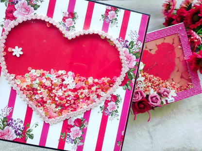A decorative heart-shaped layered craft box or gift box with a red heart cutout, pink lace trim, floral prints, and colorful sequins, shown alongside a smaller matching box on a light fabric surface with artificial flowers.