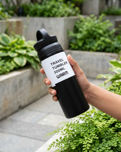 A hand holding a black travel tumbler bottle with a white label that reads 'TRAVEL TUMBLER 500ML WARM' in black text, set against a blurred green garden background.