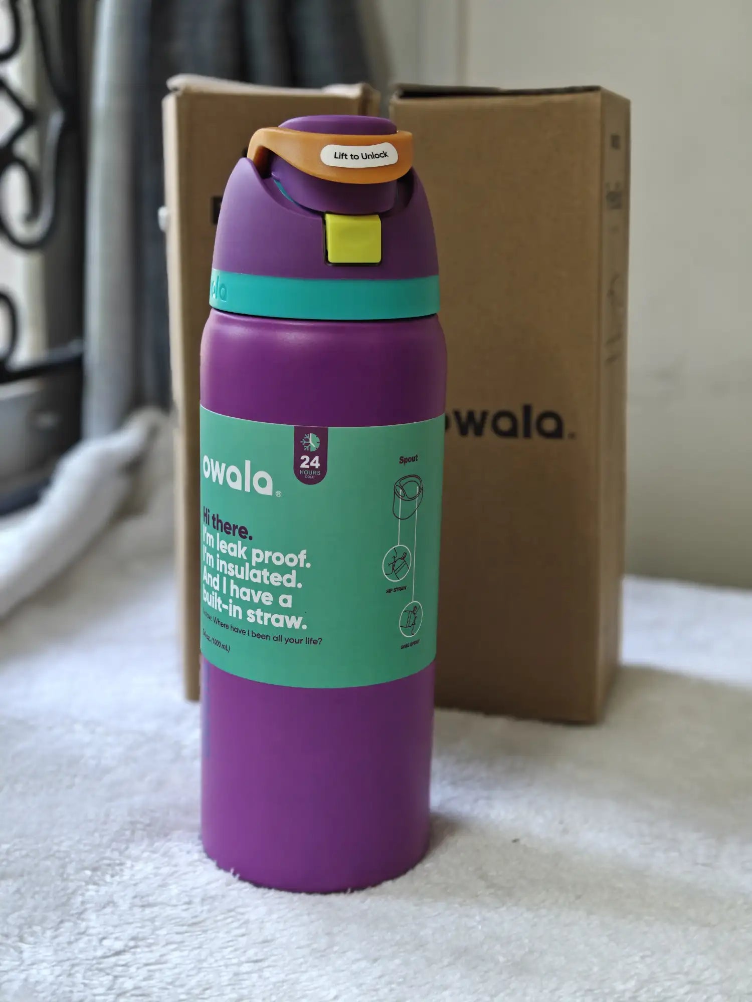 Purple water bottle with green label on a surface, with a cardboard box in the background.