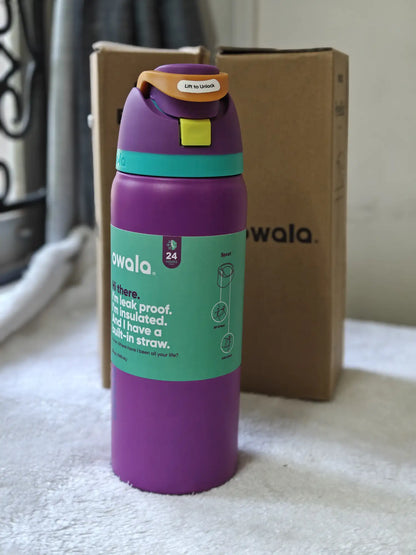 Purple water bottle with green label on a surface, with a cardboard box in the background.