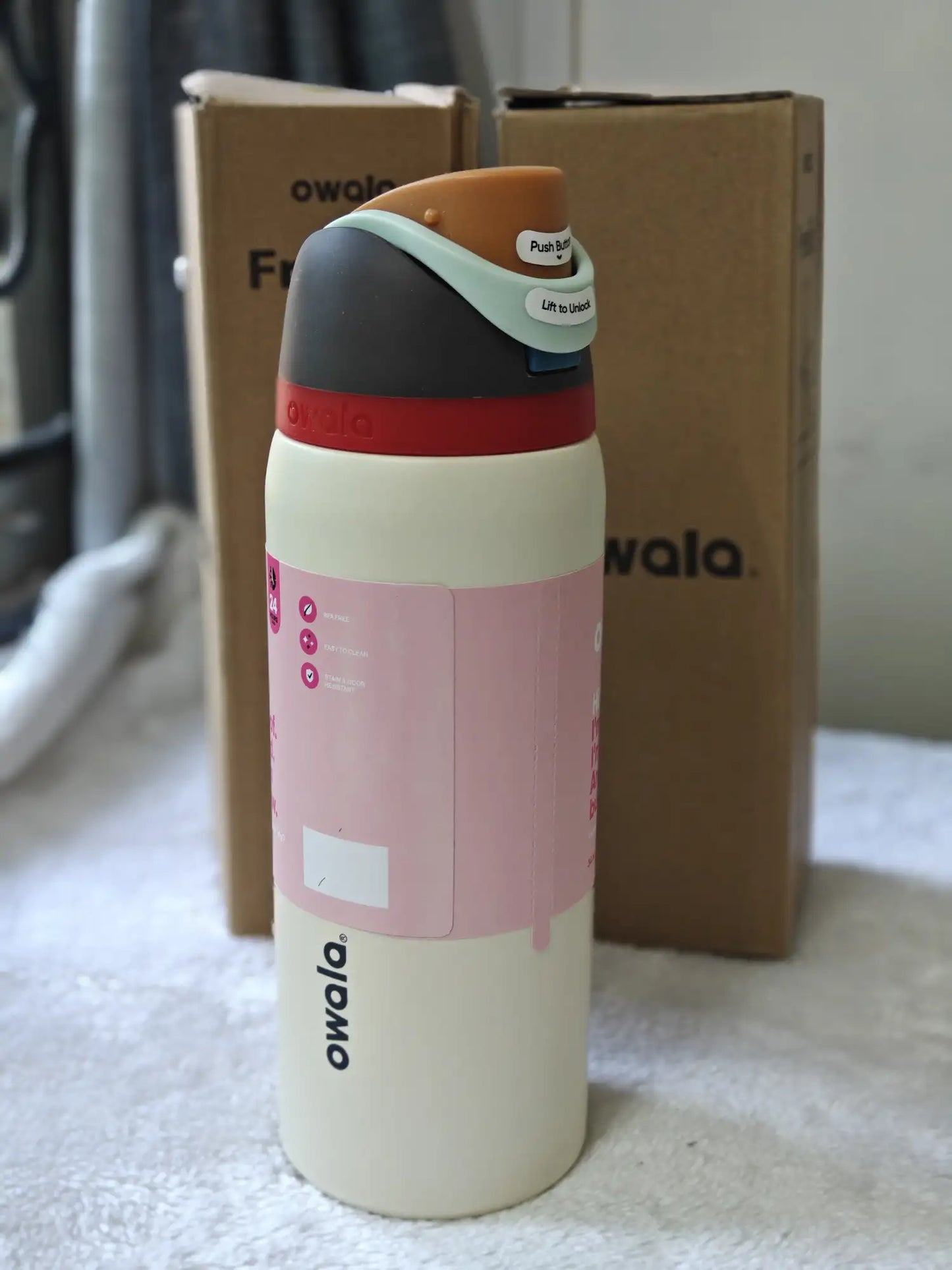 Owala water bottle with a pink label on a surface with a cardboard box in the background.