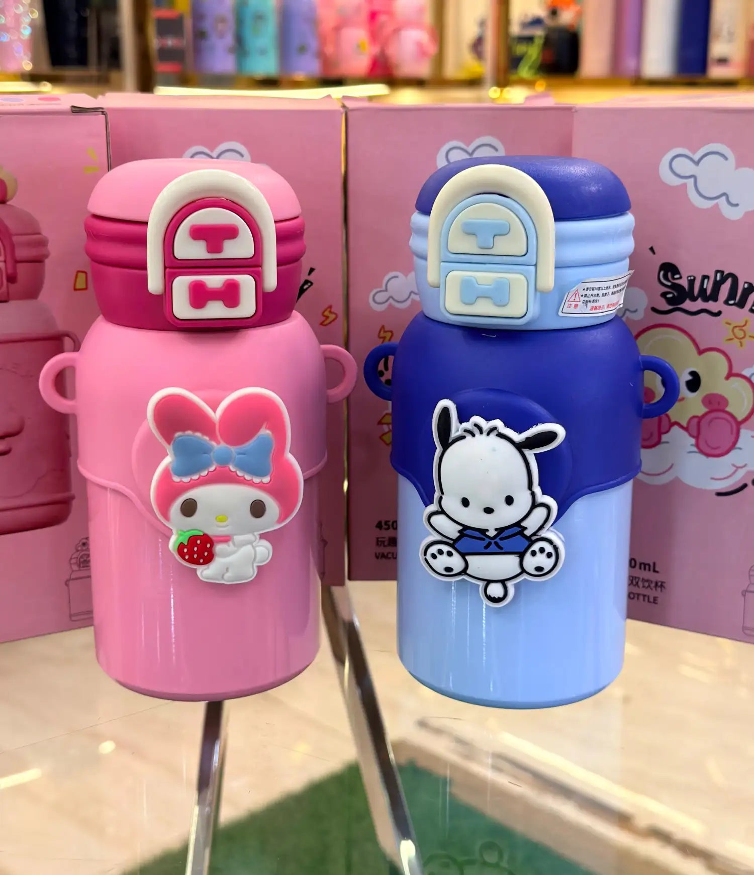 Two children's water bottles, one pink and one blue, with cartoon characters on a reflective surface.
