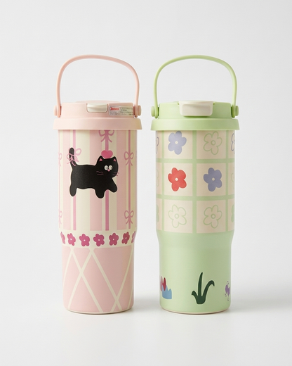 Two insulated travel tumbler cups with carry handles on a light gray studio background. The left cup is pale pink with a black cat illustration, pink bows, and floral patterns.