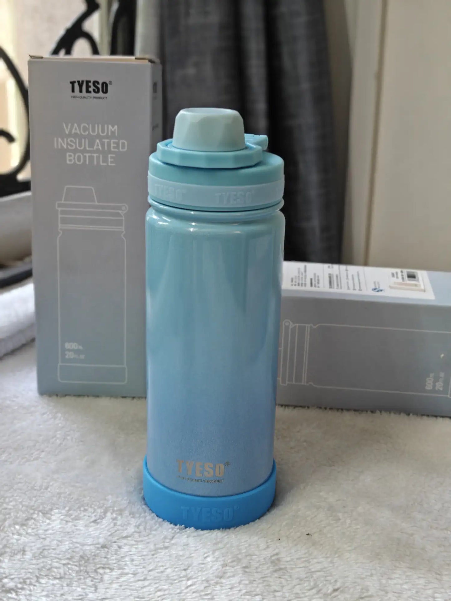 Blue TYESO vacuum insulated bottle on a white surface with packaging in the background.
