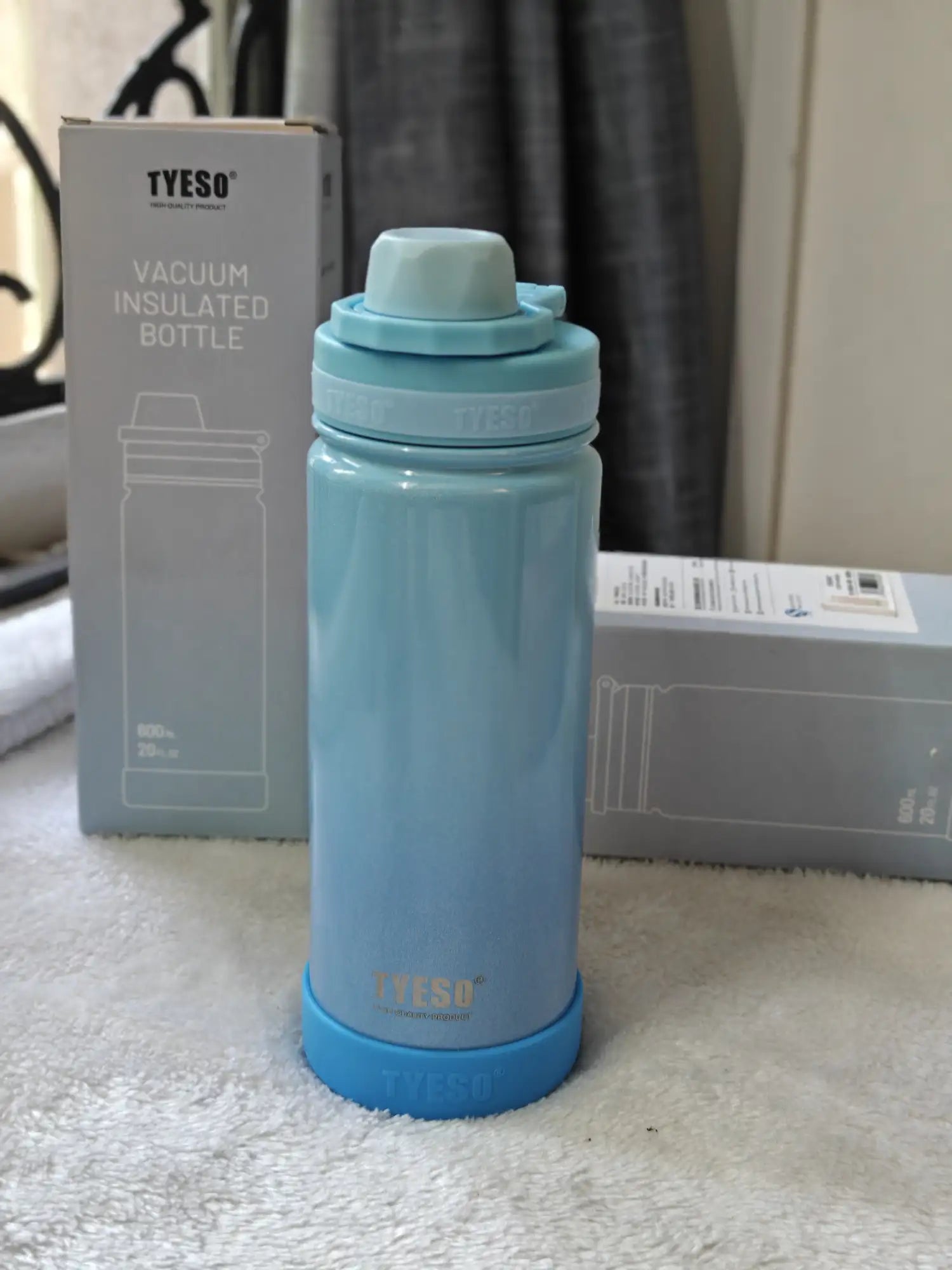 Blue TYESO vacuum insulated bottle on a white surface with packaging in the background.